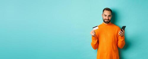 Online shopping. Handsome caucasian man in orange sweater, using credit card and mobile phone, standing over light blue background photo
