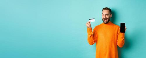 Online shopping. Handsome caucasian man showing credit card and smartphone, winking at camera, recommending something, standing over light blue background photo