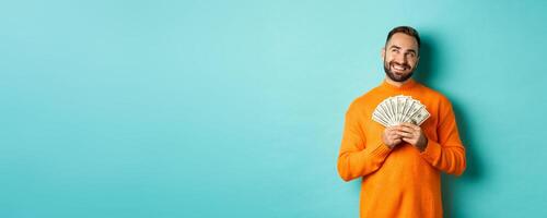 Man thinking about shopping, holding money and smiling satisfied, looking at upper left corner, standing over light blue background photo