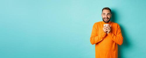 Happy young man drinking coffee, smiling and looking left with dreamy face, standing over turquoise background photo