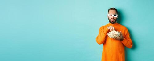 Image of intrigued man eating popcorn and watching movie in 3d, looking at tv screen, standing over blue background photo