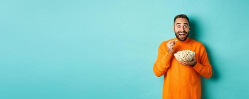 Excited young man watching interesting movie on tv screen, eating popcorn and looking amazed, blue background photo