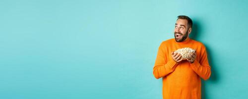 Excited and happy man watching tv and holding bowl of popcorn, looking left and smiling pleased, standing over blue background photo