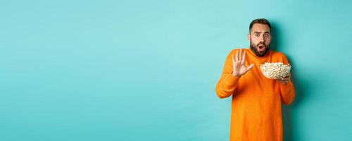 Portrait of scared guy holding bowl of popcorn and declining something, showing stop gesture, standing over blue background photo