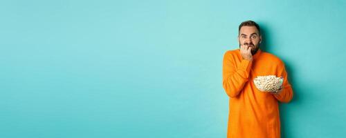 Image of scared young man watching horror movie, biting fist and looking terrified, holding bowl of popcorn, standing over turquoise background photo