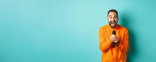 Handsome adult man perform song, singing into microphone, standing against turquoise background photo