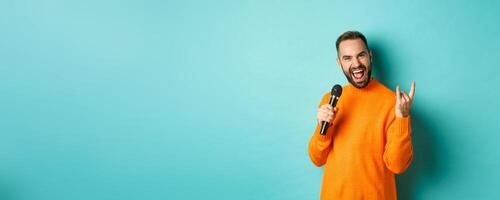 Joyful handsome man singing song in microphone, showing rock-n-roll gesture excited, standing over turquoise background photo