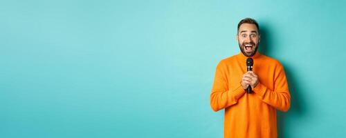 Happy adult man singing karaoke, holding microphone and looking at camera, standing in orange sweater against turquoise background photo