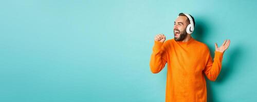 Happy man lipsync while listening music in headphones, holding invisible microphone, standing over turquoise background photo