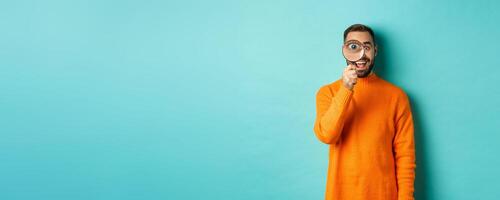 Cheerful man searching for something, looking through magnifying glass and smiling happy, standing over blue background photo