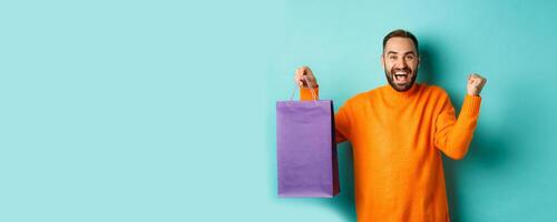 Happy man holding purple shopping bag and rejoicing, got discount and celebrating, standing over turquoise background photo
