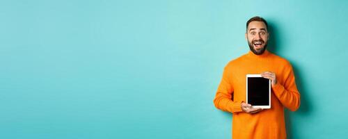 Technology. Excited adult man in orange sweater showing digital tablet screen, staring amazed at camera, standing over turquoise background photo