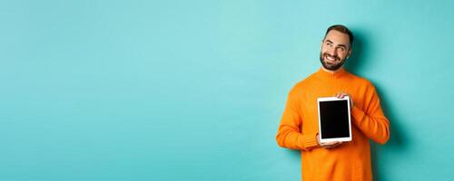 Technology. Handsome adult man showing digital tablet screen, smiling and looking at upper left corner thoughtful, imaging or daydreaming, blue background photo