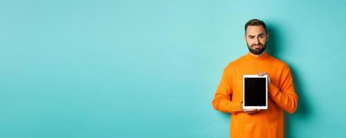 Technology. Skeptical and displeased guy showing digital tablet screen, frowning disappointed, standing over light blue background photo
