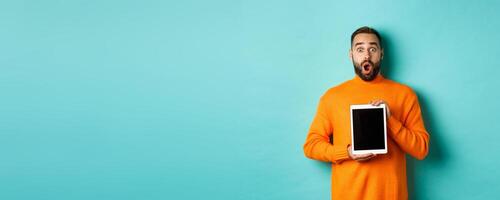 Technology. Surprised bearded man drop jaw, staring at camera and showing digital tablet screen, standing againt light blue background photo