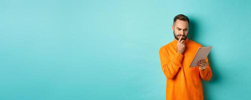Serious man pondering and looking at digital tablet screen, reading social media, standing over turquoise background photo