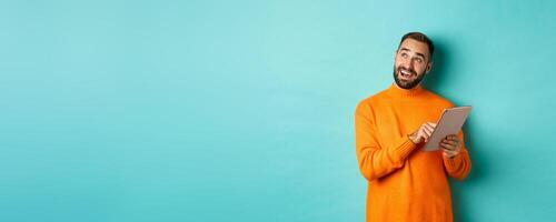 Handsome man thinking and using digital tablet, shopping online and pondering ideas, looking at upper left corner, standing over light blue background photo