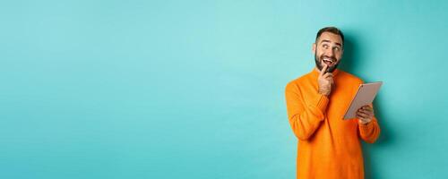 Handsome man thinking and using digital tablet, shopping online and pondering ideas, looking at upper left corner, standing over light blue background photo