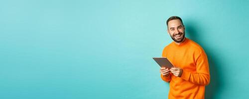 Handsome bearded man using digital tablet, laughing at camera, standing happy against light blue background photo