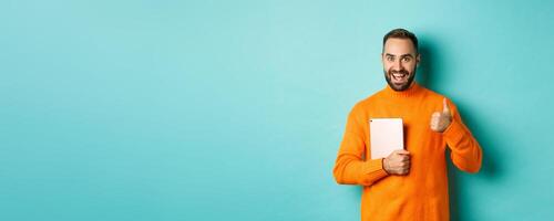 Work from home, technology concept. Handsome man holding laptop, showing thumb up, approve and like something, standing over turquoise background photo