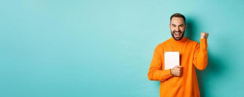 Excited adult man holding laptop and making fist pump, rejoicing of win, standing satisfied against turquoise background photo