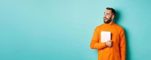 Handsome man holding laptop, looking left with surprise and amazement, standing in orange sweater against light blue background photo