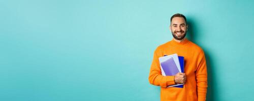 Education. Smiling bearded man holding notebooks and smiling, going on courses, standing over light blue background photo