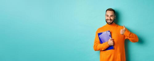 Education. Smiling bearded man holding notebooks and smiling, going on courses, showing thumbs-up in approval, like something, standing over turquoise background photo