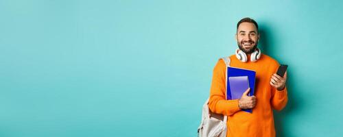 Education. Handsome male student with headphones and backpack, using smartphone and holding notebooks, smiling happy, standing over turquoise background photo