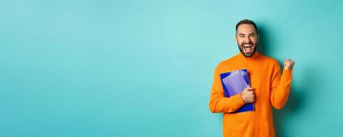 Education. Happy and satisfied man rejoicing, holding notebooks and making fist pump, shouting for joy, standing against turquoise background photo