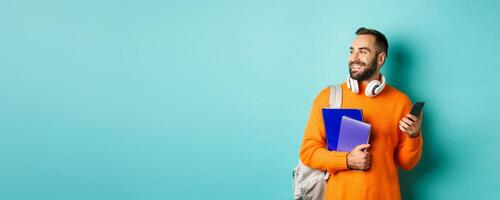 Education. Handsome male student with headphones and backpack, using mobile phone and holding notebooks, smiling happy, standing over turquoise background photo