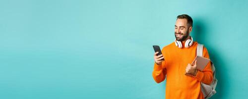 Handsome man student with headphones and backpack, holding digital tablet, reading message on mobile phone, standing against light blue background photo