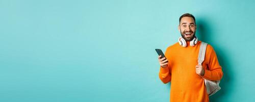 Caucasian man with headphones and backpack staring at camera amazed after reading phone notification, standing over turquoise background photo