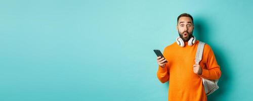 Caucasian man with headphones and backpack staring at camera surprised after reading phone message, standing over turquoise background photo