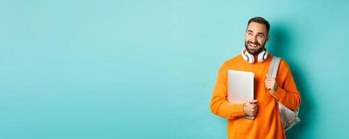 Happy man with backpack and headphones, holding laptop and smiling, looking left thoughtful, standing over turquoise background photo