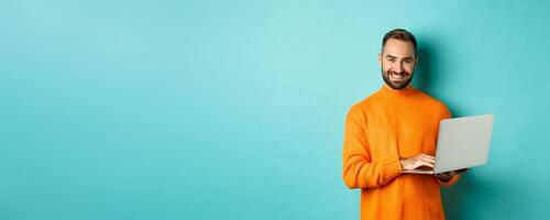 Handsome adult man freelancer working with laptop, smiling at camera, typing on computer keyboard, standing over light blue background photo
