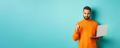 Image of adult man teaching online, showing finger while using laptop, explaining rules, standing over light blue background photo