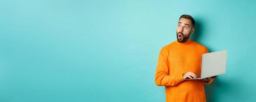 Handsome man doing online shopping, looking up thoughtful while using laptop, standing over light blue background photo