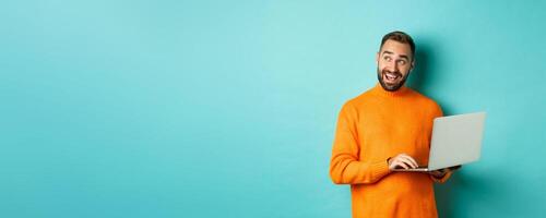Handsome man doing online shopping, looking up thoughtful while using laptop, standing over light blue background photo
