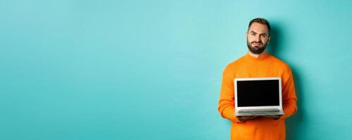 Handsome bearded man in orange sweater showing laptop screen, demonstrating promo, grimacing disappointed and upset standing over light blue background photo