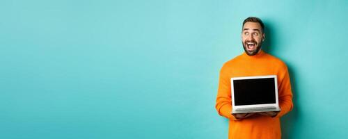 Amazed young man in orange sweater looking at upper left corner, showing laptop screen promo offer, standing over turquoise background photo
