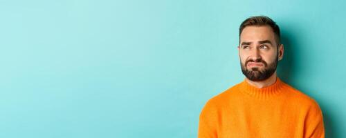 Close-up of handsome caucasian man looking left disappointed and skeptical, staring at logo, wearing orange sweater, standing against turquoise background photo