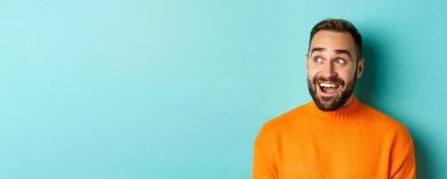 Close-up of handsome caucasian man smiling, looking left with surprised face, staring at logo, wearing orange sweater, standing against turquoise background photo