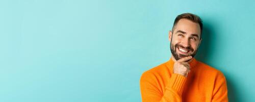 Close-up of caucasian male model having an idea, smiling and looking upper left corner thoughtful, imaging plan, standing over light blue background photo