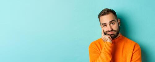 Close-up of unamused sad man looking at camera, pouting and leaning face on hand, standing over light blue background photo