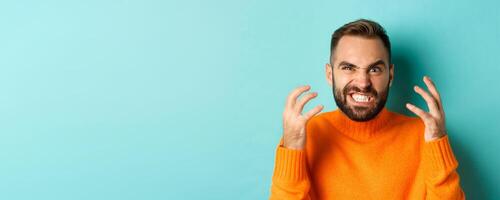 Close-up of angry caucasian man looking mad, grimacing and shaking hands, having break down, standing over light blue background photo