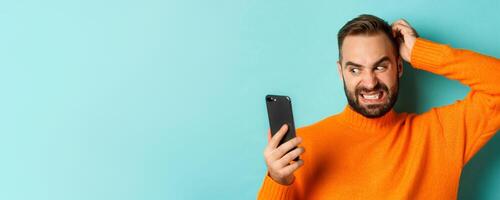 Close-up of caucasian man looking troubled and reluctant at phone screen, scratching head and grimacing confused, standing over turquoise background photo