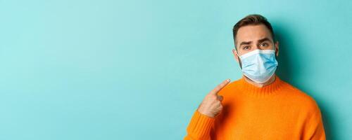 Covid-19, social distancing and quarantine concept. Upset man in complaining on face mask, pointing at it and looking bothered, standing over light blue background photo