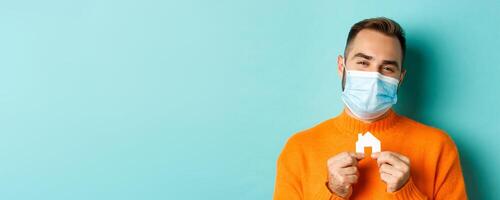 Real estate and coronavirus pandemic concept. Close-up of adult man in medical mask holding small paper house maket and smiling, searching for apartment, light blue background photo