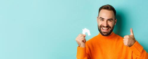 Real estate. Close-up of satisfied man showing thumb-up and small paper house maket, smiling pleased, standing over light blue background photo